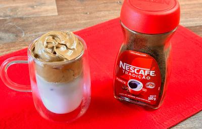 Foto aproximada da receita de Dalgona Café feito com Nescafé Tradição, servido em um xícara grande transparente ao lado de um pote de Nescafé, tudo sobre um pano vermelho em uma tábua de madeira Foto aproximada da receita de Dalgona Café feito com Nescafé Tradição, servido em um xícara grande transparente ao lado de um pote de Nescafé, tudo sobre um pano vermelho em uma tábua de madeira
