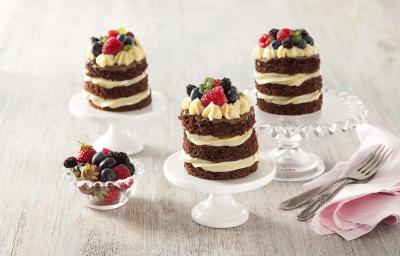 Fotografia em tons de branco e cinza de uma bancada cinza com com suportes de bolos brancos e de vidro com naked cakes. Ao lado um paninho rosa com garfos e um recipiente redondo de vidro com frutas vermelhas.