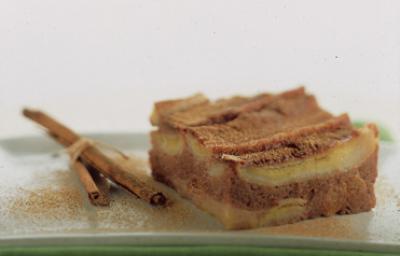 Fotografia em tons de canela e cinza, com pedaço de torta de banana com canela sobre travessa verde com ramos de canela ao lado, tudo polvilhado com canela, em fundo cinza. Fotografia em tons de canela e cinza, com pedaço de torta de banana com canela sobre travessa verde com ramos de canela ao lado, tudo polvilhado com canela, em fundo cinza.