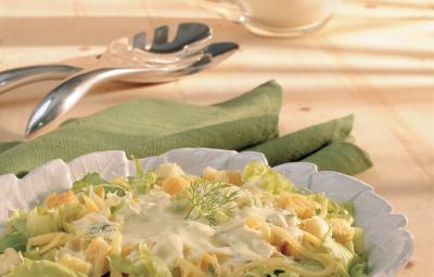 Fotografia em tons de verde em uma bancada de madeira clara, um pano verde, um pegador de salada e um prato redondo branco fundo com a salada de alface, queijo, torradinhas e iogurte dentro. Fotografia em tons de verde em uma bancada de madeira clara, um pano verde, um pegador de salada e um prato redondo branco fundo com a salada de alface, queijo, torradinhas e iogurte dentro.