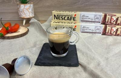 Fotografia em tons de vermelho com uma xícara de vidro ao centro. Dentro da xícara existe uma dose de café espresso. Ao fundo existe duas pimentas inteiras e duas caixas de cápsulas NESCAFE Fotografia em tons de vermelho com uma xícara de vidro ao centro. Dentro da xícara existe uma dose de café espresso. Ao fundo existe duas pimentas inteiras e duas caixas de cápsulas NESCAFE