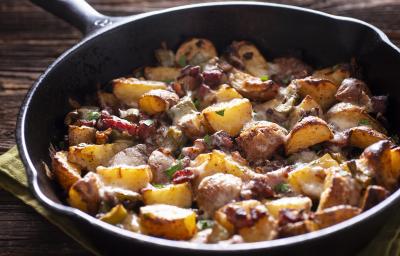 Fotografia de batatas assadas em pedaços com carne em uma panela de ferro, com salsinha por cima. A panela está apoiada em um pano verde, que está sobre uma mesa de madeira. Fotografia de batatas assadas em pedaços com carne em uma panela de ferro, com salsinha por cima. A panela está apoiada em um pano verde, que está sobre uma mesa de madeira.