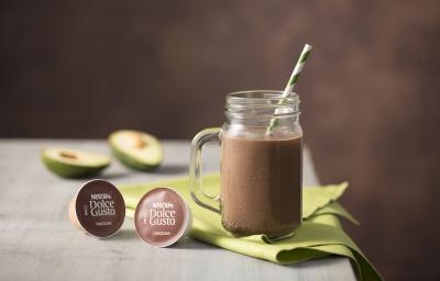 fotografia em tons de cinza e marrom de uma bancada vista de frente, contém um pano verde com uma xicara transparente com a bebida de Choccoccino Cremoso, duas capsula de Dolce Gusto, e ao fundo duas fatias de abacate