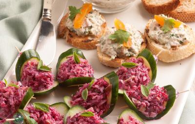 Fotografia em tons de roxo e verde em uma bancada de madeira clara com uma bandeja branca retangular grande com os canapés de pepino e beterraba. Ao lado, um potinho de vidro com patê e torradinhas. Fotografia em tons de roxo e verde em uma bancada de madeira clara com uma bandeja branca retangular grande com os canapés de pepino e beterraba. Ao lado, um potinho de vidro com patê e torradinhas.