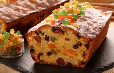 Em cima de uma mesa, bolo fofinho com frutas cristalizadas e uvas-passas, deccorado com frutas cristalizadas.