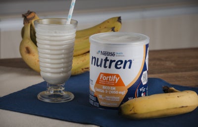 Imagem da receita de vitamina do acolhimento, uma vitamina de banana servida em um copo de vidro largo com bananas e uma lata de nutren fortify ao lado