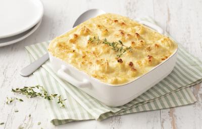Fotografia em tons de branco e verde de uma bancada de madeira branca com um paninho listrado branco e verde, sobre ele um recipiente quadrado branco com o gratinado e uma colher. Ao fundo dois pratos brancos.