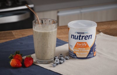 A imagem mostra a receita da vitamina fortificante servida em um copo de vidro alto e largo com um canudo dourado, ao lado há morangos, mirtilos e uma lata de nutren fortify