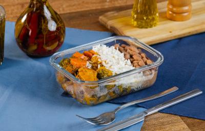 Fotografia de uma marmita retangular pequena com lombo suíno, arroz, feijão, vegetais e couve. A marmita está sobre dois panos sobrepostos de cor azul, ao lado de talheres e uma mini tábua com pimenta e azeite. Fotografia de uma marmita retangular pequena com lombo suíno, arroz, feijão, vegetais e couve. A marmita está sobre dois panos sobrepostos de cor azul, ao lado de talheres e uma mini tábua com pimenta e azeite.