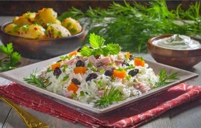 Em um prato quadrado de porcelana, há uma base de arroz soltinho, combinado com pedaços de presunto cozido, coloridos vegetais, como cenoura, uvas-passas e decorado com cebolinha picada.