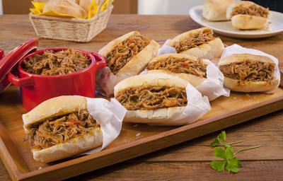 Fotografia em tons de marrom, vermelho e branco de uma bancada de madeira com uma tábua de madeira, sobre ela pães recheados com pernil desfiados dentro de saquinhos de papel brancos. Ao lado um recipiente