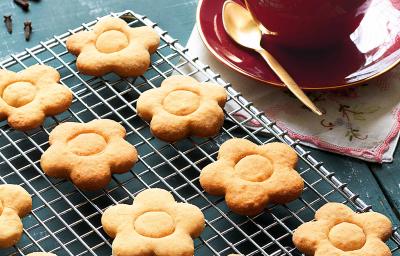 fotografia em tons de azul e bege de uma bancada azul vista de cima, contém uma grade com biscoitinhos em formato de flores e ao lado um pratinho vermelho com uma xicara vermelha que contém a bebida de chocolate branco. fotografia em tons de azul e bege de uma bancada azul vista de cima, contém uma grade com biscoitinhos em formato de flores e ao lado um pratinho vermelho com uma xicara vermelha que contém a bebida de chocolate branco.
