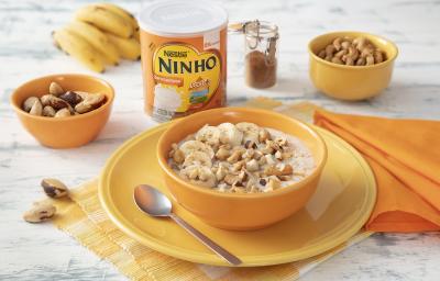 Fotografia em tons de laranja e amarelo de uma bancada de madeira, ao centro um prato amarelo e uma tigela laranja com o mingau. Ao fundo pote com castanhas, um cacho de bananas, lata de NINHO Zero Lactose e pote de canela.