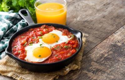 Fotografia de uma receita feita em um panela preta com molho de tomate, ovos e acompanhada de um suco de laranja