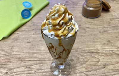 Fotografia em tons de azul e verde em uma bancada de madeira e um copo de vidro ao centro com a bebida de Milkshake e ao fundo um potinho com canela em pó.