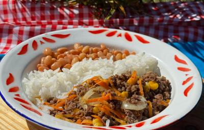Fotografia em tons claros de um prato branco com detalhes vermelhos na borda. Dentro do prato tem arroz, feijão e a carne moída com legumes. O prato está sobre um pano azul, apoiado em uma toalha vermelha e branca, quadriculada. Fotografia em tons claros de um prato branco com detalhes vermelhos na borda. Dentro do prato tem arroz, feijão e a carne moída com legumes. O prato está sobre um pano azul, apoiado em uma toalha vermelha e branca, quadriculada.