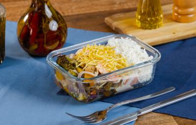 Fotografia de uma marmita retangular pequena com arroz, fricassê, batata palha, arroz e legumes assados. A marmita está sobre dois panos sobrepostos de cor azul, ao lado de talheres e uma mini tábua com pimenta e azeite. Fotografia de uma marmita retangular pequena com arroz, fricassê, batata palha, arroz e legumes assados. A marmita está sobre dois panos sobrepostos de cor azul, ao lado de talheres e uma mini tábua com pimenta e azeite.