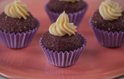Fotografia de quatro forminhas de cor roxa com um brigadeiro de açaí, e por cima de cada um tem uma cobertura de chocolate confeitado. As unidades de brigadeiro estão sobre um apoio de cor rosa. Fotografia de quatro forminhas de cor roxa com um brigadeiro de açaí, e por cima de cada um tem uma cobertura de chocolate confeitado. As unidades de brigadeiro estão sobre um apoio de cor rosa.