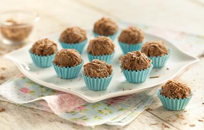 Fotografia em tons de branco, azul e marrom de uma bancada vista de frente. Panos floridos e acima um prato quadrado que contém vários brigadeiros com raspas de chocolates por cima para decorar. Fotografia em tons de branco, azul e marrom de uma bancada vista de frente. Panos floridos e acima um prato quadrado que contém vários brigadeiros com raspas de chocolates por cima para decorar.