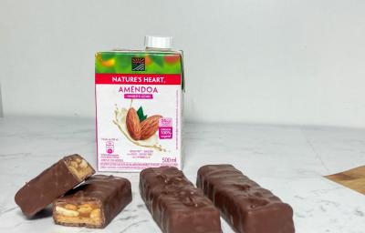 Foto em tons claros da receita de chocolate com caramelo e amendoim servida em 4 porções sobre uma mesa de mármore branca com uma embalagem de bebida vegetal de amêndoa nature's heart ao fundo Foto em tons claros da receita de chocolate com caramelo e amendoim servida em 4 porções sobre uma mesa de mármore branca com uma embalagem de bebida vegetal de amêndoa nature's heart ao fundo