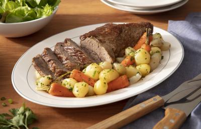 Fotografia em tons de verde em uma bancada de madeira clara, um prato oval branco com a maminha fatiada e legumes em cima dele. Ao fundo, um recipiente com salada, talheres de churrasco.