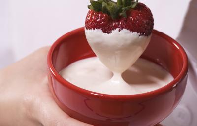 Fotografia de uma chef segurando um morango coberto de fondue de chocolate branco, que está dentro de um recipiente de vidro na cor vermelha. Fotografia de uma chef segurando um morango coberto de fondue de chocolate branco, que está dentro de um recipiente de vidro na cor vermelha.