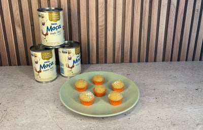 Docinhos claros cobertos com açúcar em forminhas laranja, dispostos em prato verde com Leite Moça ao fundo.