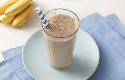 Fotografia em tons de azul e marrom de uma bancada com um paninho azul dobrado, um prato azul e um copo com a bebida de chocolate e um canudo. Ao fundo um cacho de bananas.