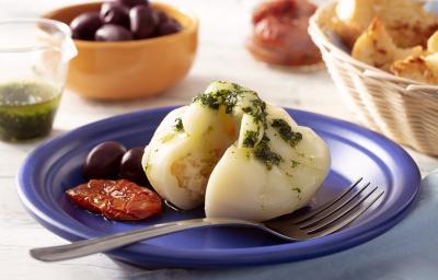Fotografia em tons de branco em uma bancada de madeira de cor branca. Ao centro, um prato azul contendo a burrata com um garfo ao lado. Ao fundo, está uma cesta com pães e tomates secos. Fotografia em tons de branco em uma bancada de madeira de cor branca. Ao centro, um prato azul contendo a burrata com um garfo ao lado. Ao fundo, está uma cesta com pães e tomates secos.