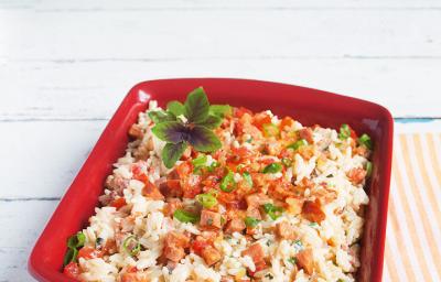 Fotografia em tons de vermelho e branco de uma bancada branca com uma pano listrado laranja e branco, uma travessa vermelha retangular com o arroz, pedaços de tomate e linguiça e manjericão. Fotografia em tons de vermelho e branco de uma bancada branca com uma pano listrado laranja e branco, uma travessa vermelha retangular com o arroz, pedaços de tomate e linguiça e manjericão.