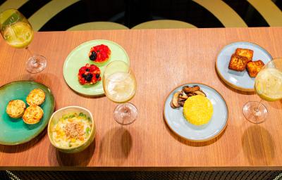 Fotografia de uma mesa de madeira com um brunch com cuscuz, quiche de alho poró, cheesecake, salada de frutas e suco de abacaxi com hortelã. As comidas estão em pratos coloridos, e o suco estão em taças, na imagem são três.