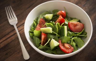 Fotografia de um recipiente fundo branco com uma salada de folhas, pepino e tomates. Ao lado do recipiente, um garfo, que está sobre uma mesa de madeira escura.
