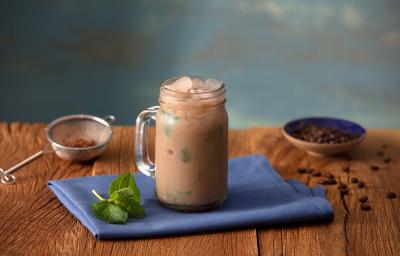 Fotografia em tons de azul em uma bancada de madeira escura, um pano azul, uma caneca alta de vidro com o cappuccino gelado de cacau, banana e hortelã. Ao lado, uma peneira com o cacau e um potinho com algumas gotas de chocolate.