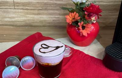 Fotografia em tons de vermelho em uma bancada de madeira, um pano vermelho e uma xícara com café e espuma rosa decorado com um coração com calda de chocolate ao centro. Ao fundo, um vaso vermelho com flores e em formato de coração. Fotografia em tons de vermelho em uma bancada de madeira, um pano vermelho e uma xícara com café e espuma rosa decorado com um coração com calda de chocolate ao centro. Ao fundo, um vaso vermelho com flores e em formato de coração.