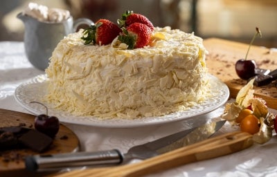 O mesmo bolo aparece inteiro, coberto com raspas de chocolate branco e decorado com morangos no topo. Está sobre um prato branco, cercado por utensílios e frutas, pronto para ser servido em uma mesa posta. O mesmo bolo aparece inteiro, coberto com raspas de chocolate branco e decorado com morangos no topo. Está sobre um prato branco, cercado por utensílios e frutas, pronto para ser servido em uma mesa posta.