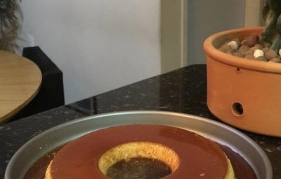 Fotografia em tons de marrom em uma bancada de madeira de cor preta. Ao centro, uma forma de alumínio contendo o pudim. Fotografia em tons de marrom em uma bancada de madeira de cor preta. Ao centro, uma forma de alumínio contendo o pudim.