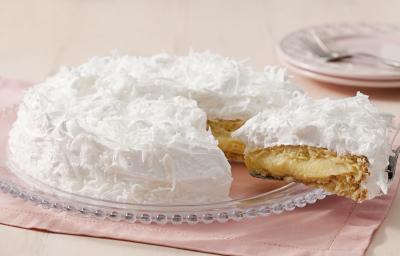 Fotografia de um bolo coberto com marshmallow branco e raspada de coco em cima. Dentro do bolo creme e massa brancos. O prato está em cima de um guardanapo de papel rosa claro.