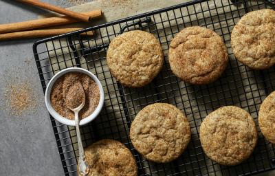 Fotografia vista de cima de sete cookies com açúcar e canela sobre uma grade preta. Ao lado dos cookies e por cima da grade, tem um pequeno pote de vidro branco com açúcar e canela, e uma colher de chá apoiada sobre ele. Fotografia vista de cima de sete cookies com açúcar e canela sobre uma grade preta. Ao lado dos cookies e por cima da grade, tem um pequeno pote de vidro branco com açúcar e canela, e uma colher de chá apoiada sobre ele.
