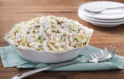 Fotografia em tons de azul em uma bancada de madeira escura, um pano azul com um recipiente redondo branco em cima dele e a salada de macarrão com peito de frango dentro dele. Ao lado, uma colher de servir macarrão. Fotografia em tons de azul em uma bancada de madeira escura, um pano azul com um recipiente redondo branco em cima dele e a salada de macarrão com peito de frango dentro dele. Ao lado, uma colher de servir macarrão.