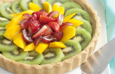 Fotografia em tons de verde, branco, amarelo e vermelho, com torta de frutas sobre porcelana branca com uma espátula e guardanapo verde ao lado, tudo sobre bancada em tons de branco e cinza. Fotografia em tons de verde, branco, amarelo e vermelho, com torta de frutas sobre porcelana branca com uma espátula e guardanapo verde ao lado, tudo sobre bancada em tons de branco e cinza.
