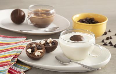 Fotografia em tons de branco e amarelo de uma bancada cinza, ao centro um prato com uma xícara com leite e o chocobomb e ao lado um chocobomb aberto, mostrando a camada de chocolate e o recheio.