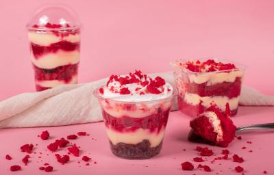 Fotografia em tons de rosa com alguns bolos de pote de red velvet ao centro. Dentro de cada pote, existe camadas de creme branco alternadas com bolo red velvet Fotografia em tons de rosa com alguns bolos de pote de red velvet ao centro. Dentro de cada pote, existe camadas de creme branco alternadas com bolo red velvet