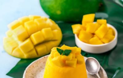 Fotografia de um flan de manga e calda de carambola, com pequenos pedaços de manga por cima. Ao lado da sobremesa, uma colher pequena que está sobre um miniprato de cerâmica branco com pontos pretos. Ao fundo, manga em pedaços e uma inteira. Fotografia de um flan de manga e calda de carambola, com pequenos pedaços de manga por cima. Ao lado da sobremesa, uma colher pequena que está sobre um miniprato de cerâmica branco com pontos pretos. Ao fundo, manga em pedaços e uma inteira.