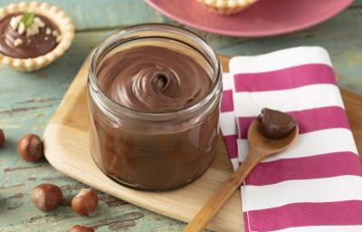 Fotografia em tons de roxo em uma bancada de madeira esverdeada, um pano branco com listras roxas, uma tábua de madeira, um potinho cilíndrico de vidro com o creme de avelã dentro dele. Ao fundo, mini tortinhas com o creme e avelãs espalhadas na mesa.