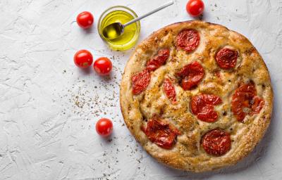 Fotografia vista de cima de uma focaccia com tomate cereja assado, sal grosso e azeite. Ao lado da focaccia tem um recipiente de vidro pequeno com azeite, ao lado alguns tomates cereja, sobre uma mesa branca com texturas.