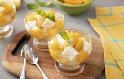 Foto da receita de Gelatina de Verão. Observa-se três potes individuais de vidro com as gelatina em quadrados e frutas amarelas picadas sobre uma tábua de madeira. Ao lado dos potes, duas colherinhas acompanham. Foto da receita de Gelatina de Verão. Observa-se três potes individuais de vidro com as gelatina em quadrados e frutas amarelas picadas sobre uma tábua de madeira. Ao lado dos potes, duas colherinhas acompanham.