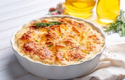Em cima de uma mesa, refratário com batata gratinada, decorado com cebolinha alecrim.