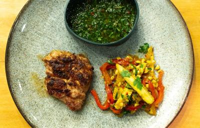 Fotografia em tons de amarelo com um prato azul ao centro. Em cima do prato existe uma receita de churrasco de timo acompanhado de chimichurri e pimentões e mini-milhos tostados. Fotografia em tons de amarelo com um prato azul ao centro. Em cima do prato existe uma receita de churrasco de timo acompanhado de chimichurri e pimentões e mini-milhos tostados.