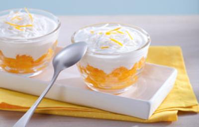 Fotografia em tons de amarelo em uma bancada de madeira clara, um pano amarelo, um recipiente branco retangular com duas taças de vidro com a sobremesa de laranja com creme de coco dentro delas. Fotografia em tons de amarelo em uma bancada de madeira clara, um pano amarelo, um recipiente branco retangular com duas taças de vidro com a sobremesa de laranja com creme de coco dentro delas.