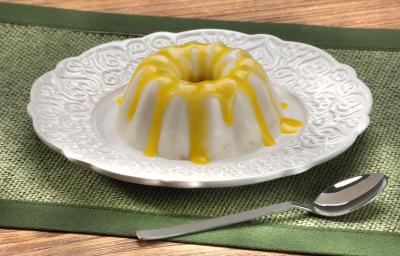 Fotografia em tons de amarelo e verde em uma bancada de madeira com uma toalha verde, um prato redondo branco raso e o flan de coco com calda de pequi em cima dele. Ao lado, uma colher. Fotografia em tons de amarelo e verde em uma bancada de madeira com uma toalha verde, um prato redondo branco raso e o flan de coco com calda de pequi em cima dele. Ao lado, uma colher.